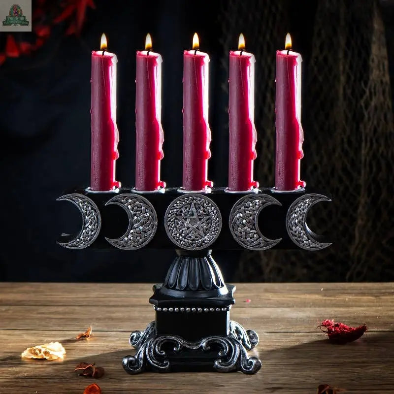 The Triple Moon Candleholder, crafted from black cold cast resin, holds five red candles; each base features silver crescent moons and a central pentagram, and it stands elegantly amid scattered dried petals.