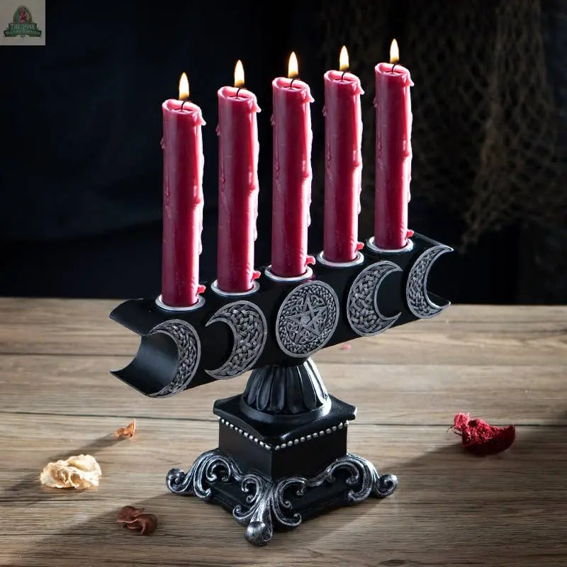 The Triple Moon Candleholder, crafted from black cold cast resin and decorated with silver crescent moons and a pentagram, holds five lit red candles. It sits on a wooden table surrounded by scattered dried flowers.