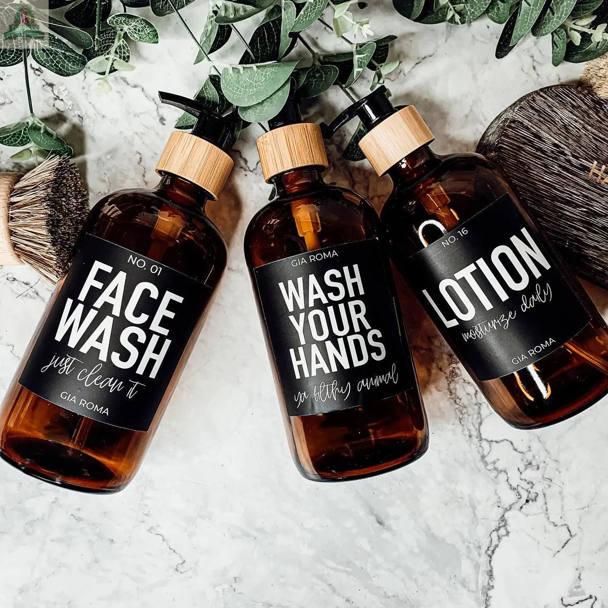 Three 16oz Glass Amber bottles with black labels reading FACE WASH, WASH YOUR HANDS, and LOTION sit on a marble bathroom tray, surrounded by green leaves and brushes. Perfect refill bottles to add style to your space.