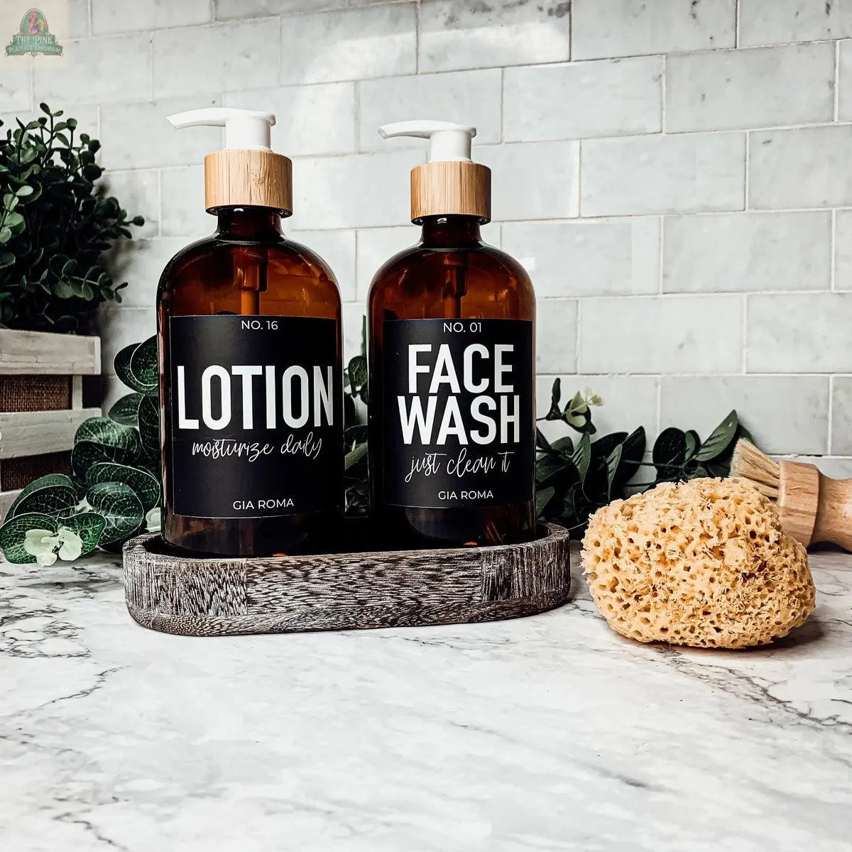 Two 16oz Glass Amber | Black Label bottles, one for lotion and one for face wash, sit on a tray with greenery, a yellow sponge, and a wooden brush on a white marble bathroom counter—ideal as stylish refill soap bottles.