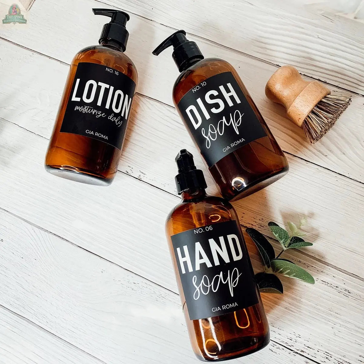 Three 16oz Glass Amber | Black Label bottles labeled Lotion, Dish Soap, and Hand Soap are arranged on a white surface with a scrub brush and greenery—ideal refill bottles for any bathroom countertop dispenser.