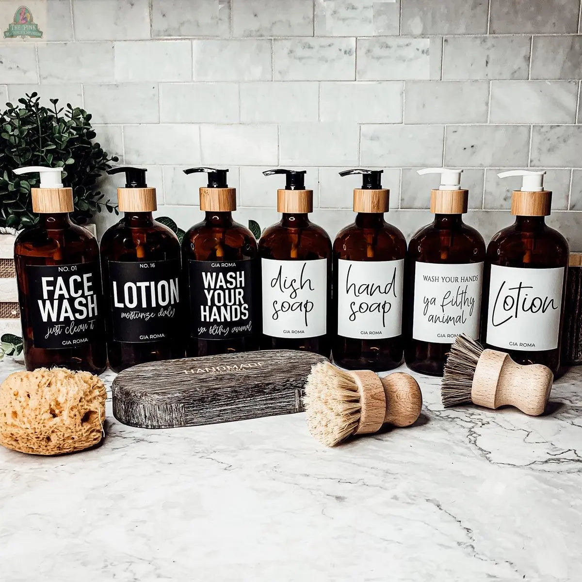 A row of 16oz Glass Amber | Black Label bottles labeled for face wash, lotion, hand wash, dish soap, and hand soap, paired with wooden cleaning brushes and a sponge—ideal refill bottles for an organized kitchen countertop.