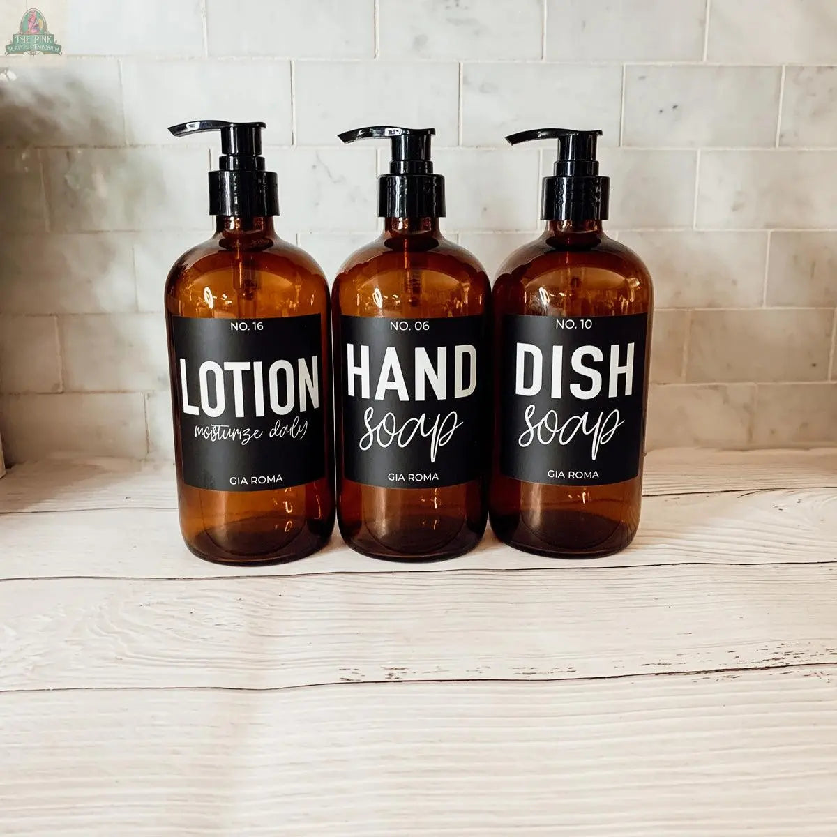 Three 16oz Glass Amber | Black Label bottles labeled Lotion, Hand Soap, and Dish Soap stand side by side on a white wooden surface with a tiled backsplash—elegant refill bottles perfect for a bathroom countertop dispenser setup.