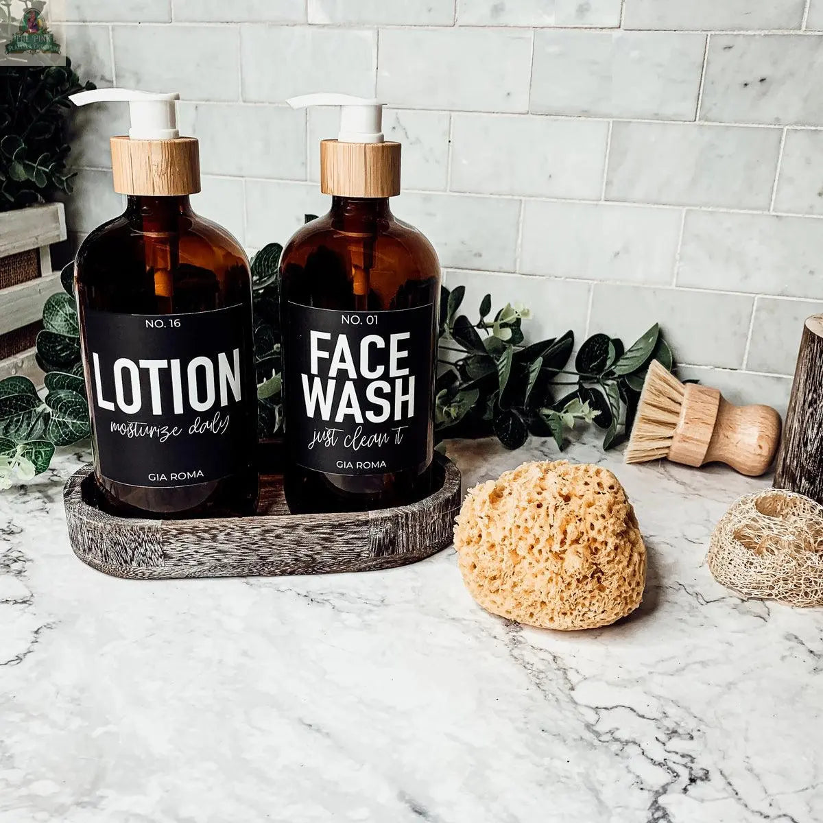 Two 16oz Glass Amber | Black Label bottles labeled Lotion and Face Wash sit in a tray on a bathroom counter, with a natural sponge, wooden brush, greenery, and tile backsplash in the background.