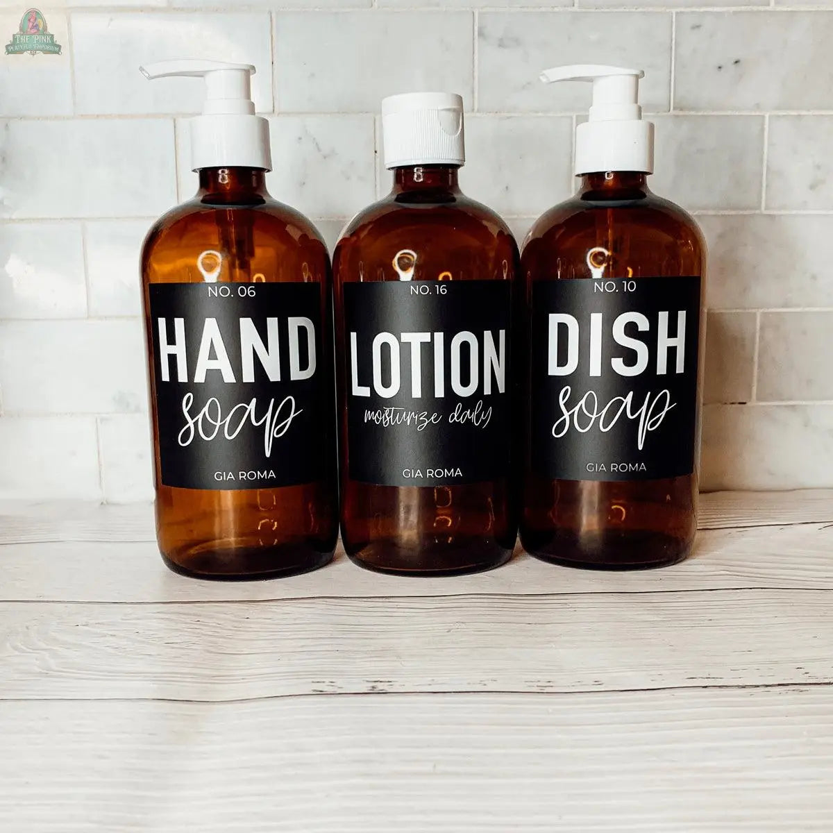 Three 16oz Glass Amber bottles with black labels—HAND SOAP, LOTION, and DISH SOAP—are lined up on a white wooden surface, creating stylish refill options for any bathroom countertop dispenser.
