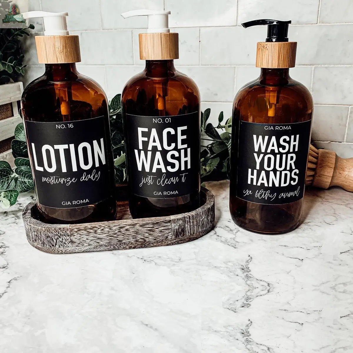 Three 16oz Glass Amber | Black Label bottles, labeled Lotion, Face Wash, and Wash Your Hands, sit on a marble counter as stylish dispensers, complementing a white tile backsplash and green foliage decor.