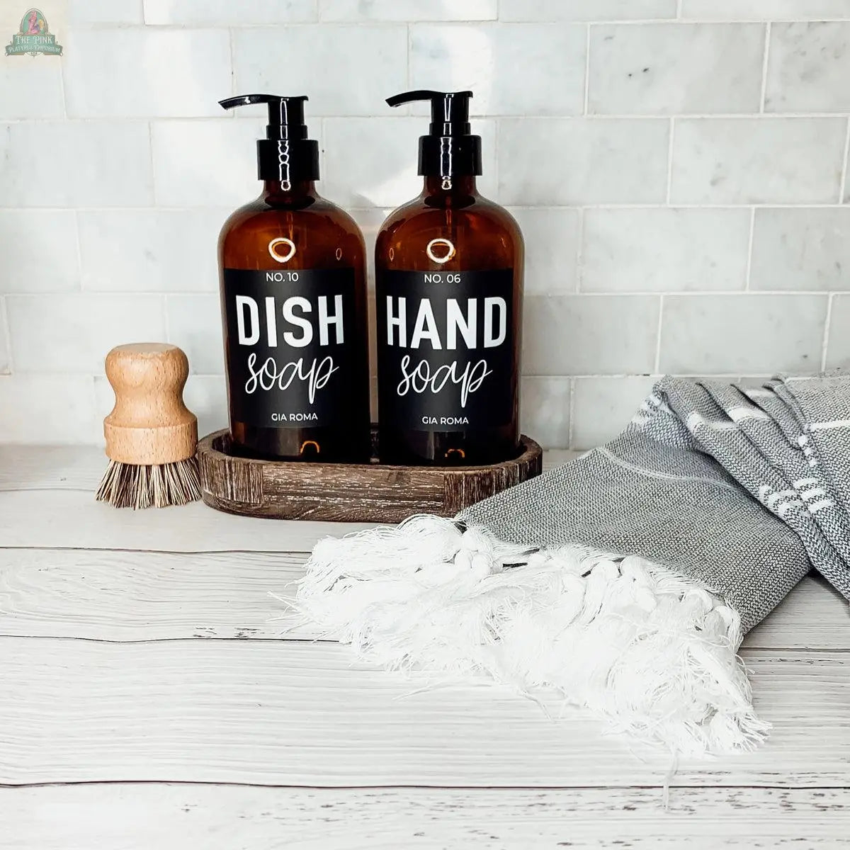 Two 16oz Glass Amber | Black Label bottles, labeled Dish Soap and Hand Soap, sit in a wooden tray on a white counter with a dish brush and a gray-white fringed towel. Perfect as stylish refill bottles for any space, with white tile backsplash behind.