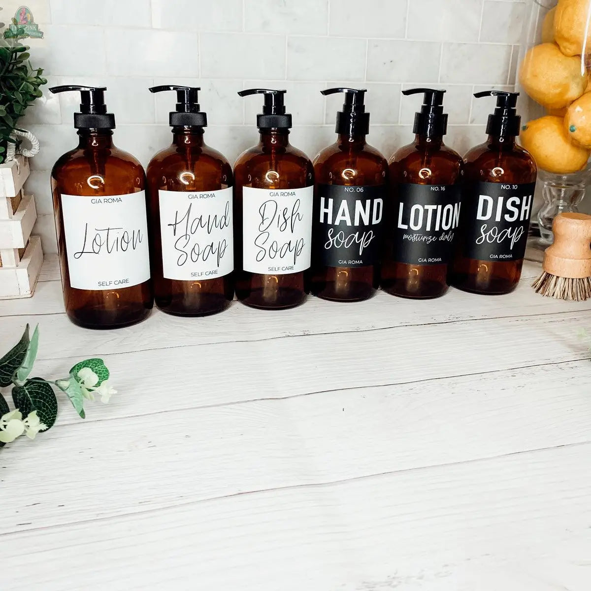 Seven 16oz Glass Amber bottles with black labels—“Lotion,” “Hand Soap,” and “Dish Soap”—are arranged on a white countertop with plants, lemons, and a wooden brush, serving as stylish refill bottles or bathroom dispensers.