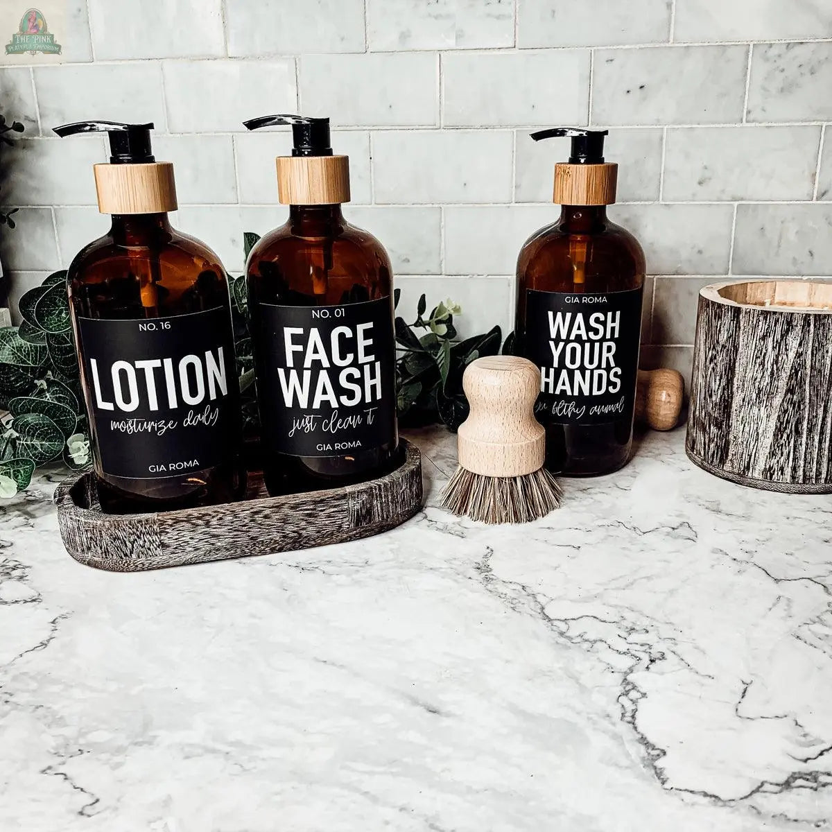 Three 16oz Glass Amber | Black Label soap bottles sit on a marble bathroom countertop alongside a scrub brush, a wooden container, and green leaves in the background.