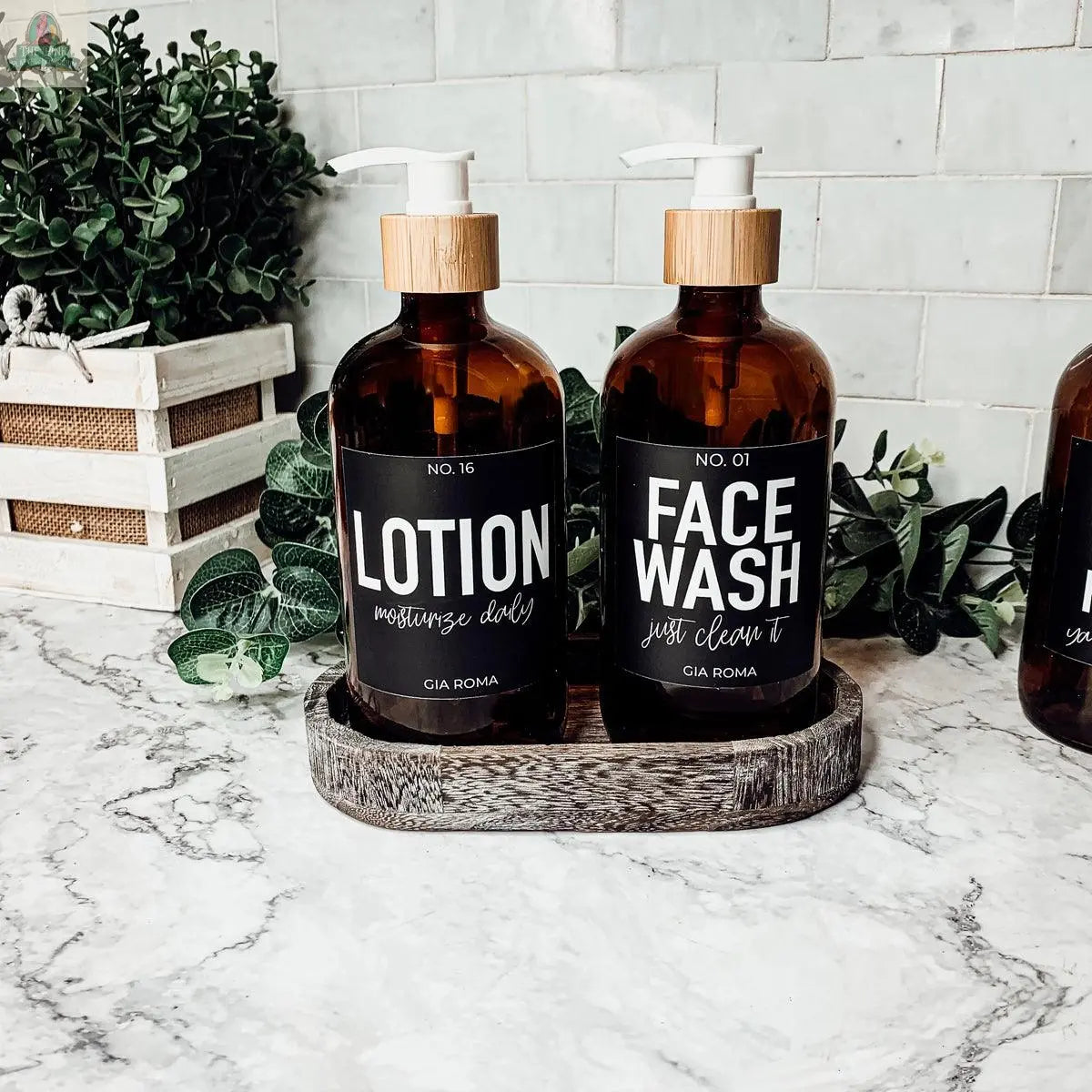 Two 16oz Glass Amber bottles with black labels, ideal for Lotion and Face Wash, sit on a marble countertop tray with green plants and a white basket—perfect as stylish refill bottles for your bathroom dispenser.
