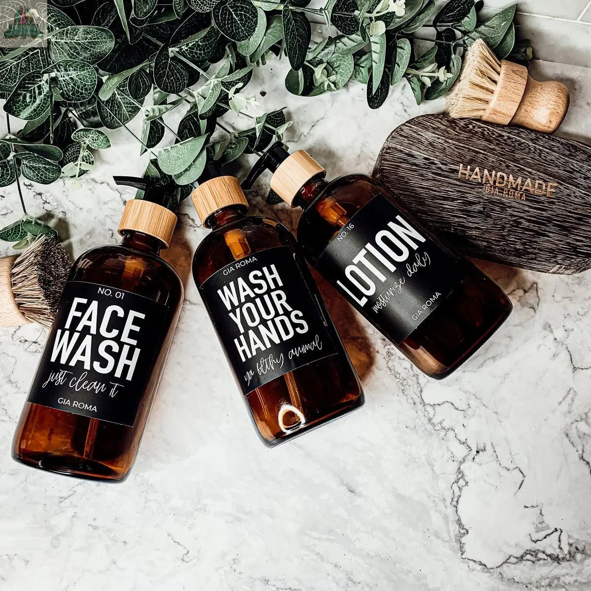 Three 16oz Glass Amber | Black Label bottles labeled face wash, hand wash, and lotion rest on a marble surface with greenery and bath accessories, adding a clean, natural touch to your bathroom countertop dispenser.