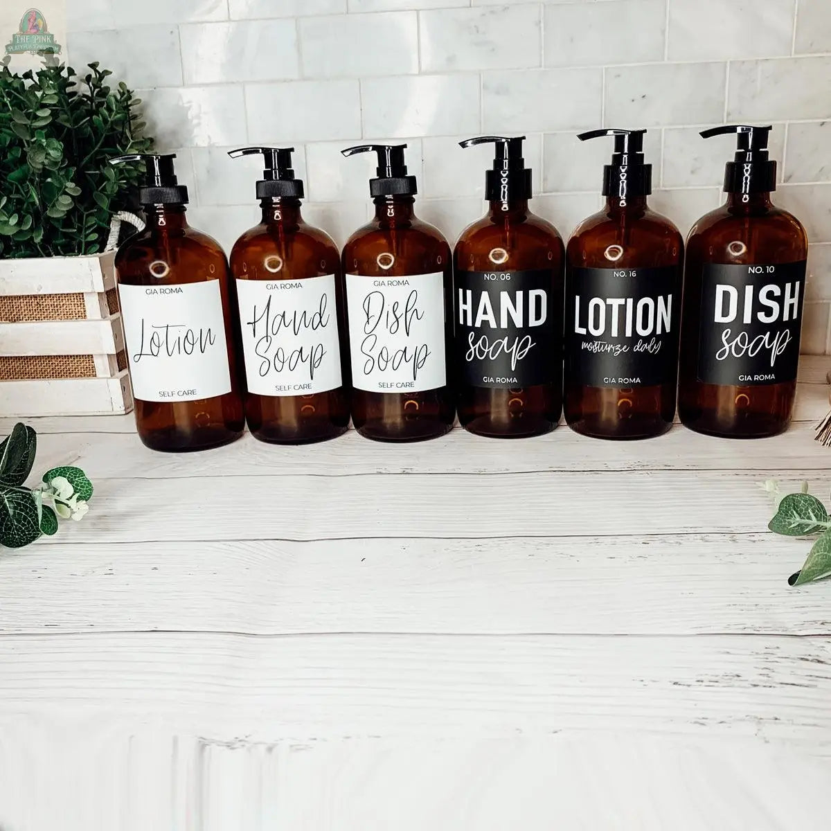 Six 16oz Glass Amber | Black Label bottles are lined up on a bathroom countertop, labeled Hand Soap, Lotion, or Dish Soap, with refill bottles among green plants and a white tiled wall in the background.