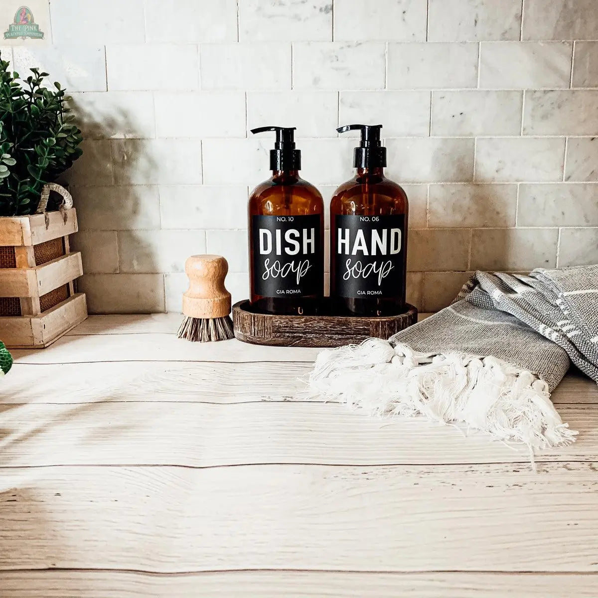 Two 16oz Glass Amber | Black Label bottles labeled Dish Soap and Hand Soap sit on a wooden tray atop a light wood counter, serving as stylish refill bottles beside a scrub brush, crate plant, and towel against the tiled backsplash.