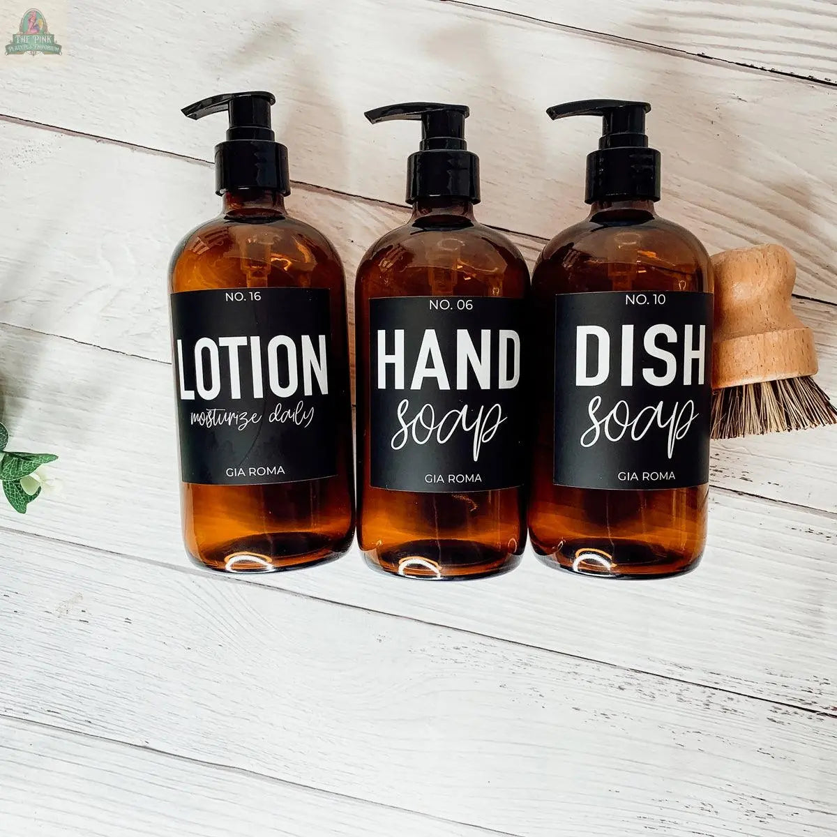 Three 16oz Glass Amber | Black Label bottles, ideal as lotion, hand soap, or dish soap dispensers, sit on a white wooden surface with a wooden scrub brush and greenery—perfect for refills or bathroom countertops.