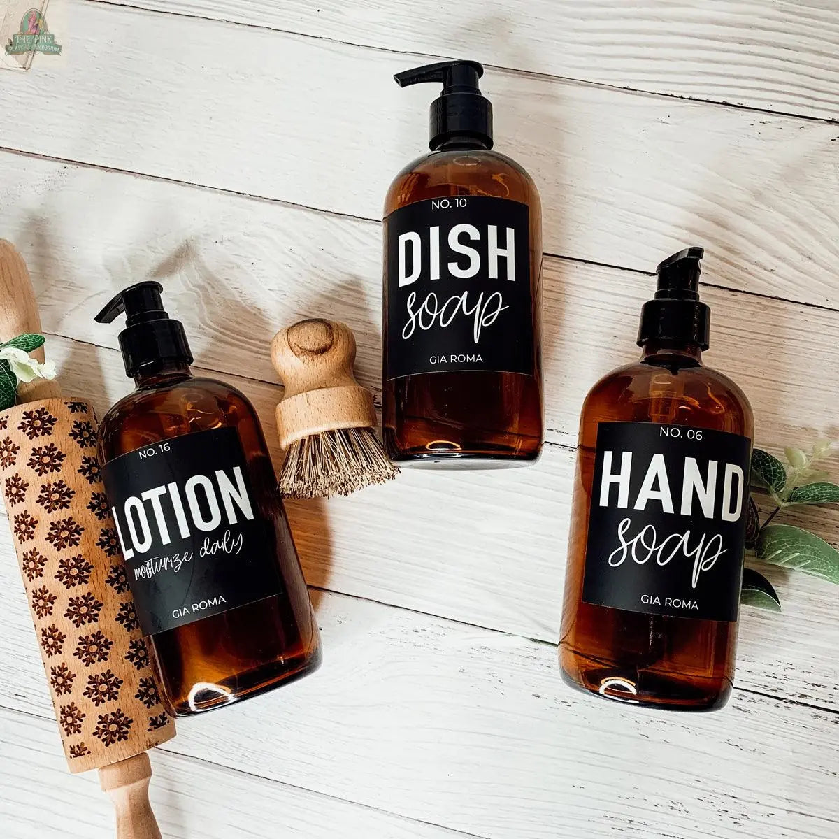 Three 16oz Glass Amber | Black Label bottles labeled “Dish Soap,” “Hand Soap,” and “Lotion” sit on a white surface with a scrub brush, rolling pin, and plant—perfect as stylish refill bottles or bathroom countertop dispensers.