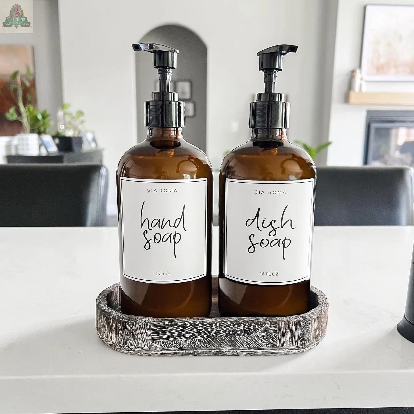 Two 16oz Glass Amber | White Label dispensers sit side by side in a rustic wooden tray on a kitchen counter, their stylish waterproof labels adding durability, with a modern kitchen and plants in the background.