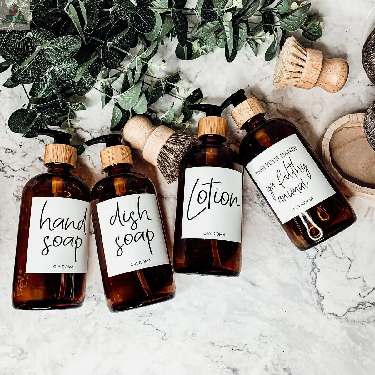 Four 16oz Glass Amber | White Label bottles for hand soap, dish soap, lotion, and a quote are arranged on a marble surface with a soap dispenser, wooden cleaning brushes, and greenery in the background.