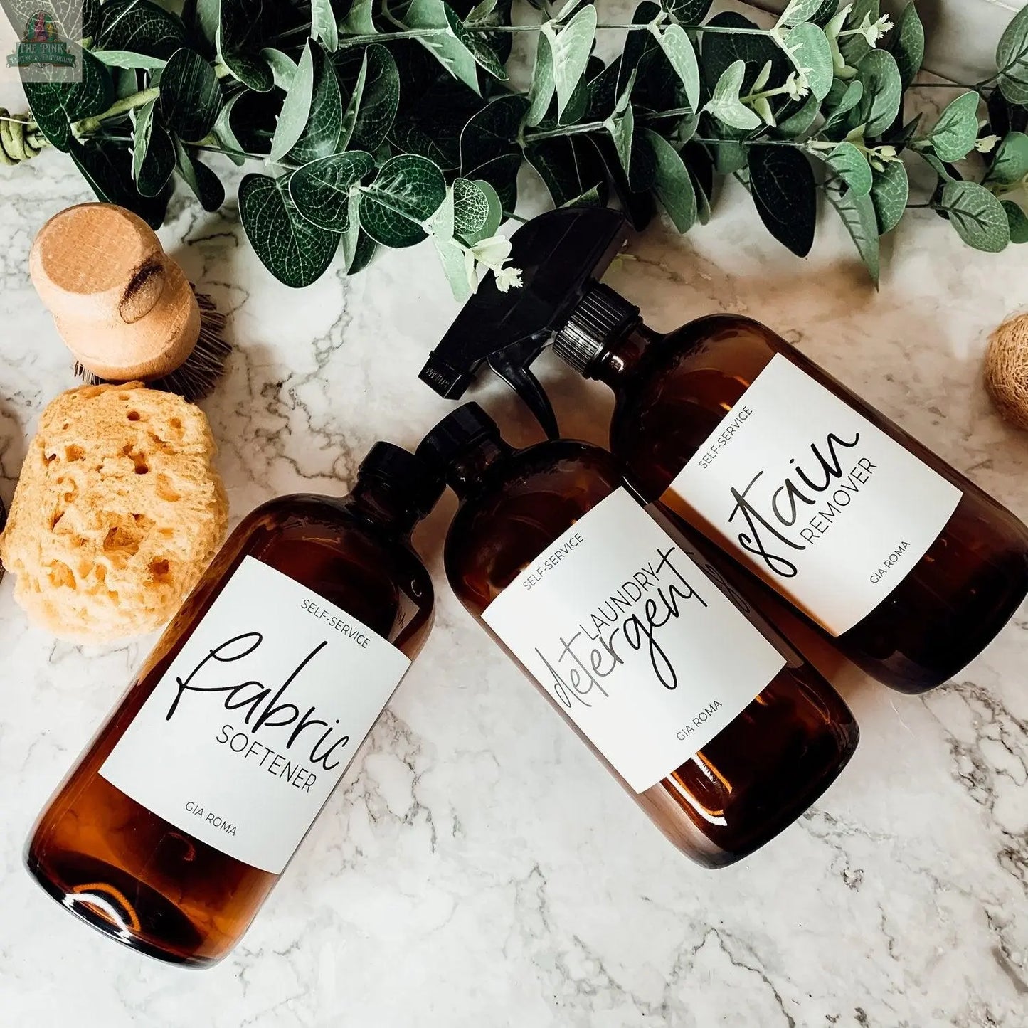Three 16oz Glass Amber | White Label bottles—labeled Fabric Softener, Laundry Detergent, and Stain Remover—sit on a marble surface with a natural sponge, wooden brush, and green leafy branches.