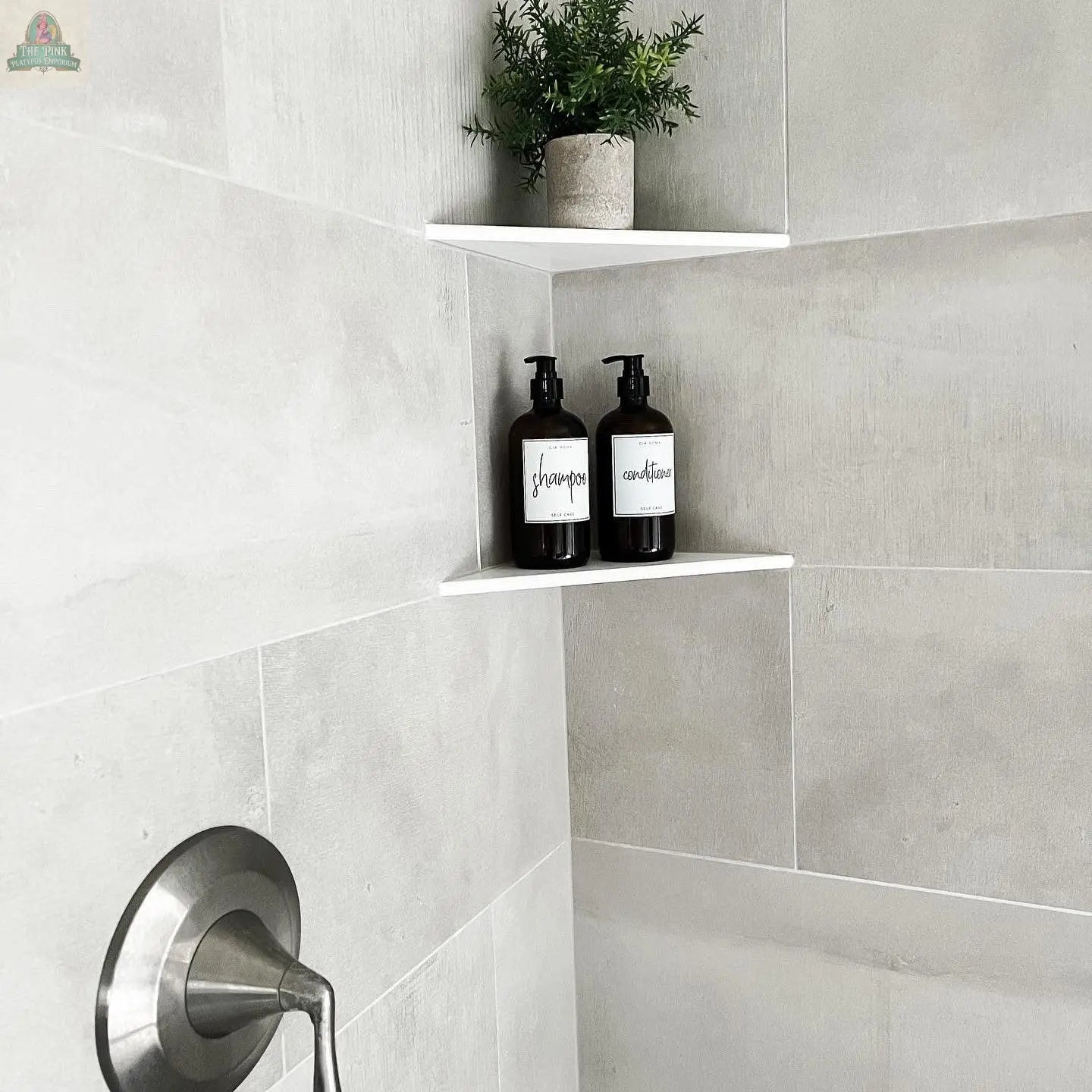 White tiled shower corner with two wall shelves: one with a potted plant, the other with 16oz Glass Amber | White Label bottles for shampoo and conditioner. A silver shower handle is below.