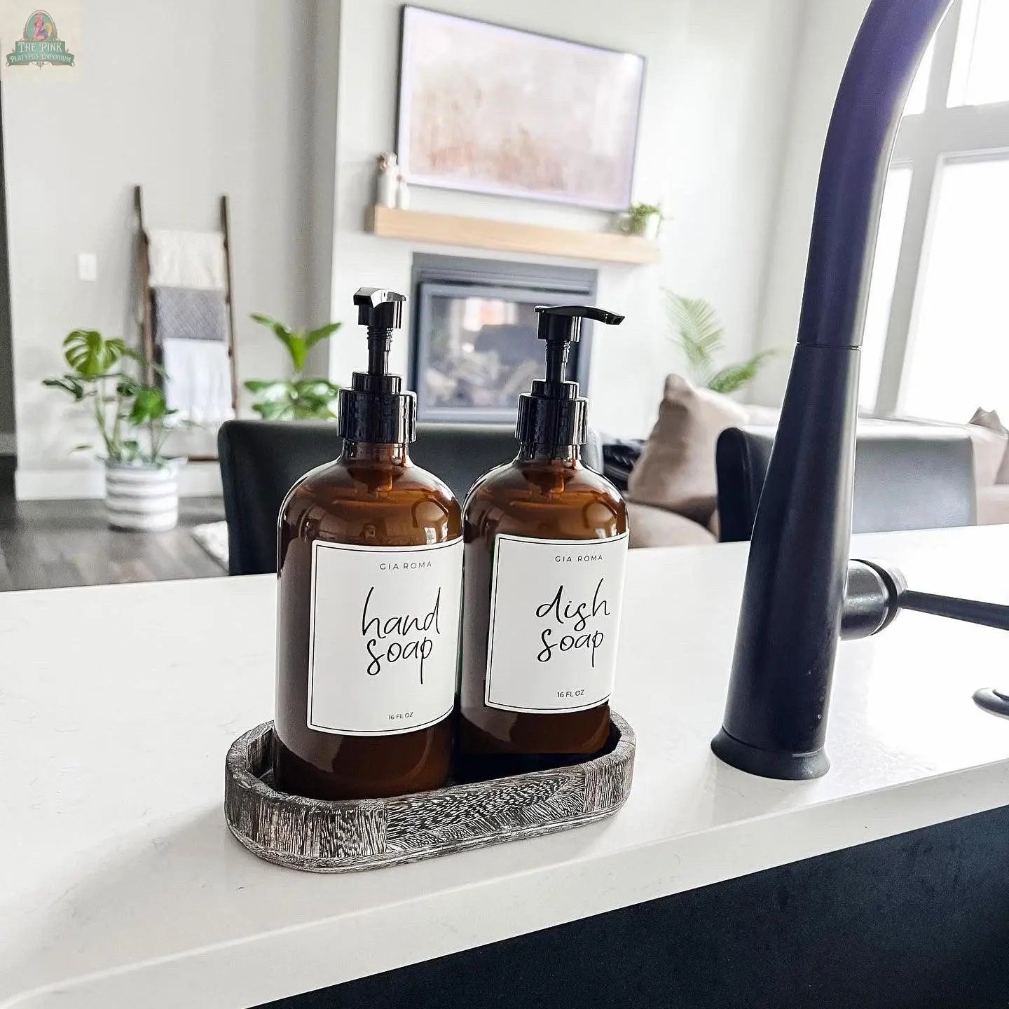 Two 16oz Glass Amber | White Label bottles for hand soap and dish soap rest in a small tray on a white kitchen counter, with a cozy living room featuring a couch, fireplace, and lush plants in the background.