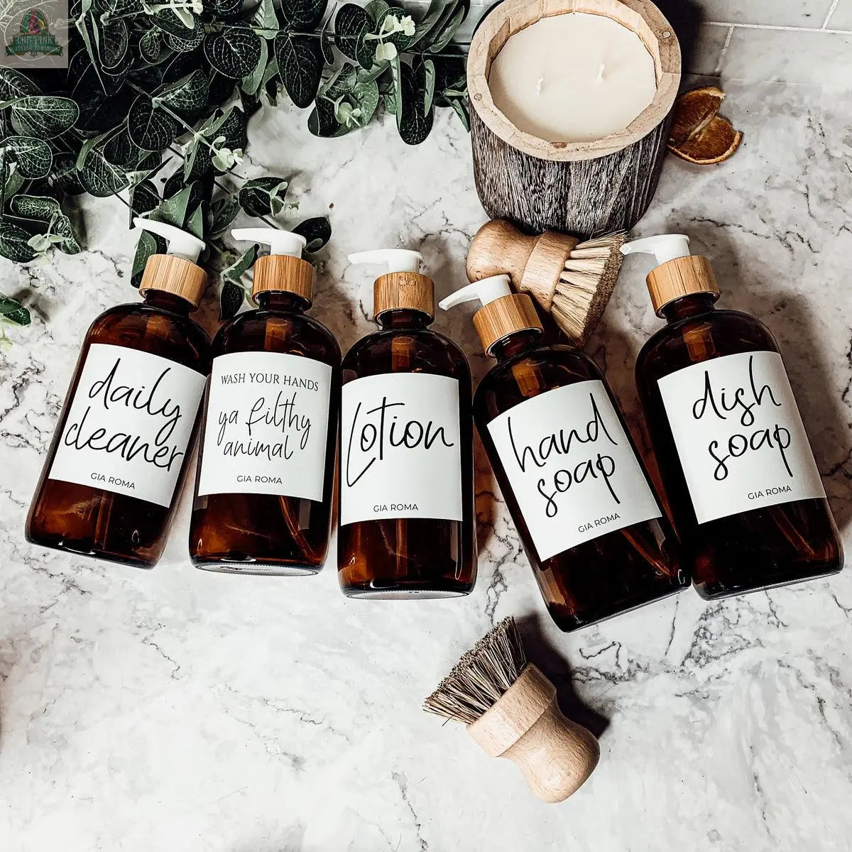 Five 16oz Glass Amber | White Label bottles with pump dispensers and waterproof labels—daily cleaner, wash your hands ya filthy animal, lotion, hand soap, and dish soap—sit on a marble counter beside brushes, a candle, and green leaves.