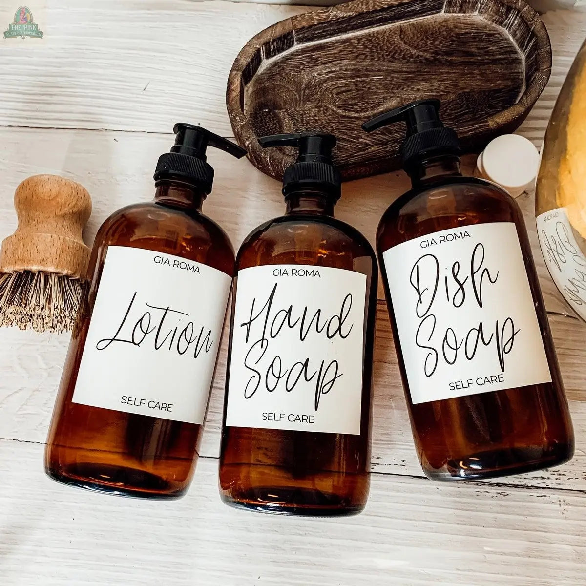 Three 16oz Glass Amber | White Label bottles for Lotion, Hand Soap, and Dish Soap are arranged on a white wooden surface, each with waterproof labels, alongside a wooden brush and soap dish.
