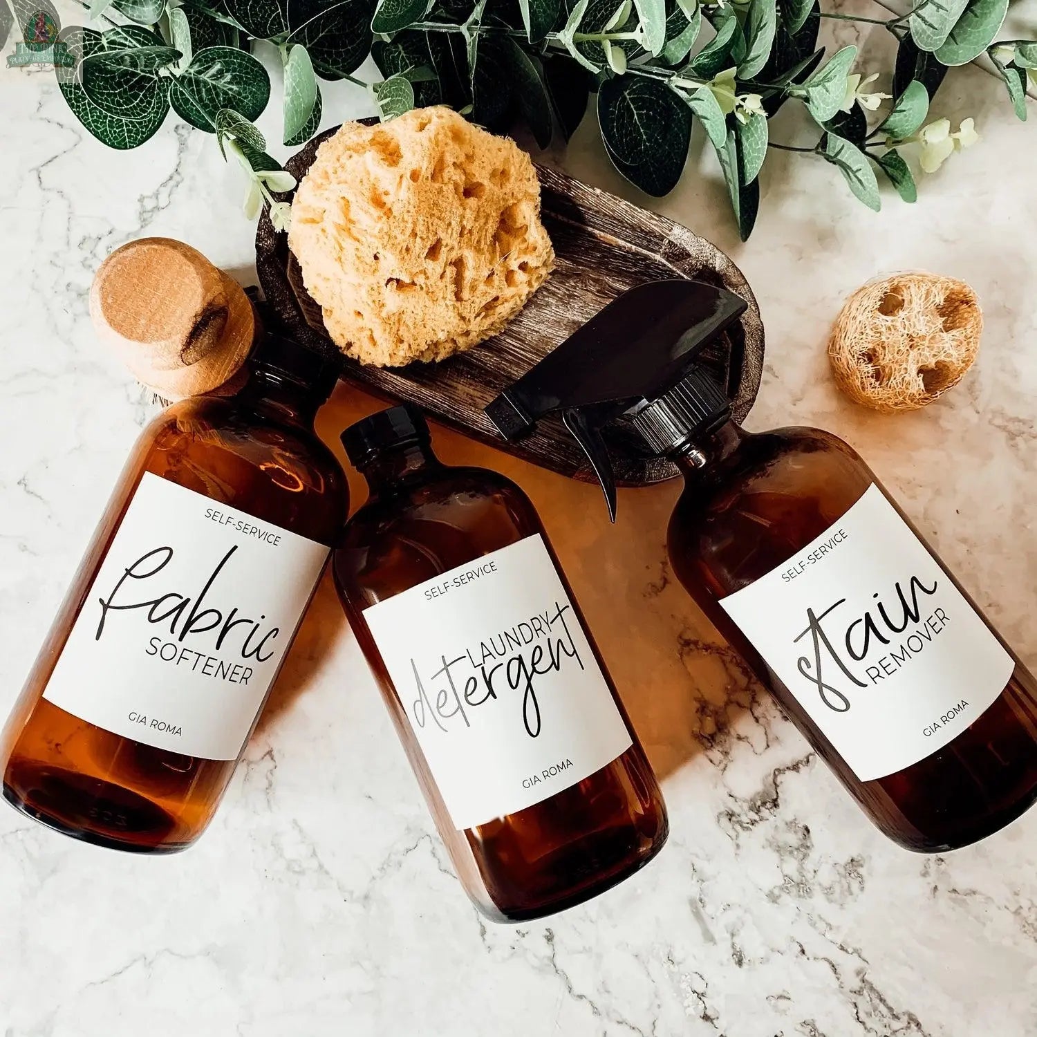 Three 16oz Glass Amber | White Label bottles are arranged on a marble surface, labeled Fabric Softener, Laundry Detergent, and Stain Remover. They’re paired with a sponge, small brush, round loofah, and green leaves above them.