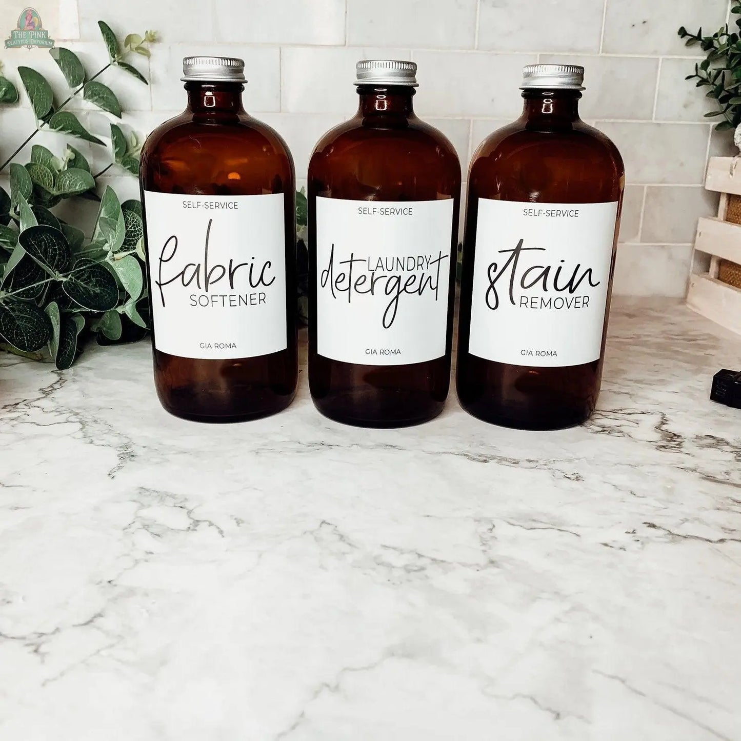 Three 16oz Glass Amber | White Label bottles labeled fabric softener, laundry detergent, and stain remover rest on a marble countertop with green plants and a tile backsplash, resembling elegant soap dispensers.