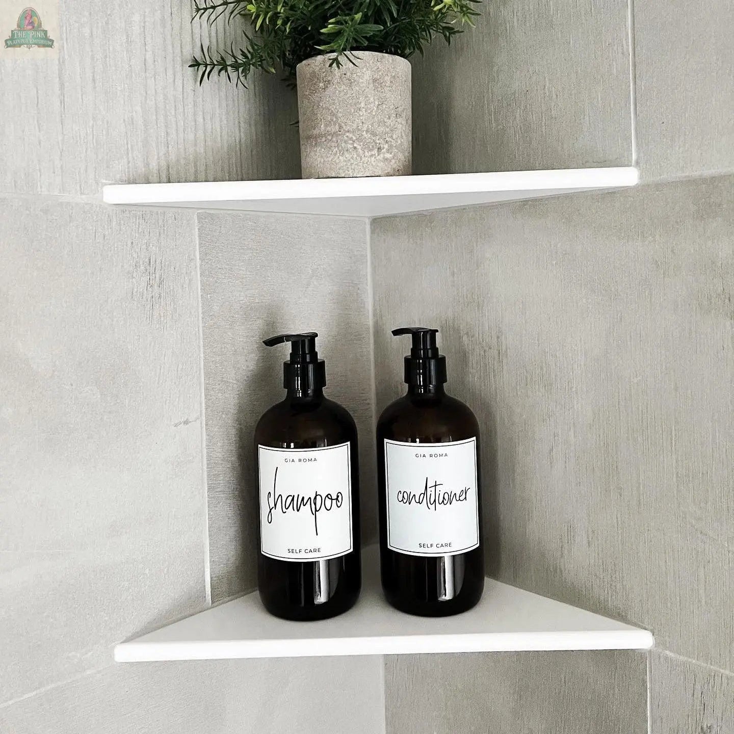 Two 16oz Glass Amber | White Label bottles, marked shampoo and conditioner with waterproof labels, sit on a white corner shelf in a tiled bathroom. A small potted plant is placed on the shelf above them.