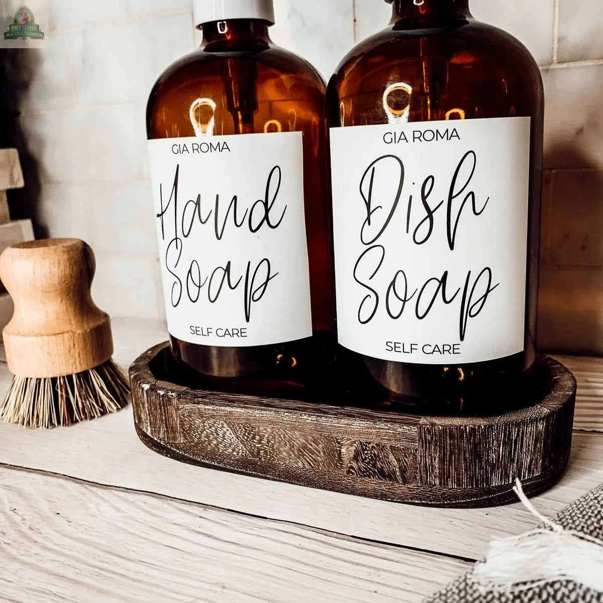 Two 16oz Glass Amber | White Label bottles labeled Hand Soap and Dish Soap sit on a wooden tray atop a light wood countertop. A scrub brush is nearby, with a cloth partially visible in the foreground.