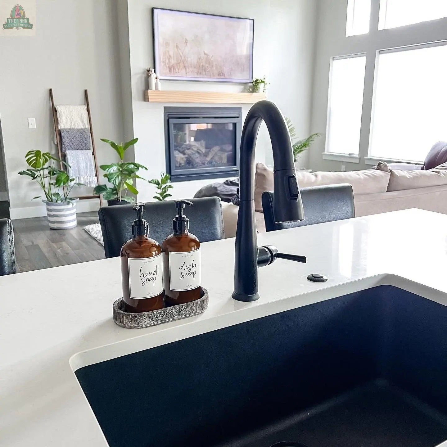 Modern kitchen with a black faucet and sink, white countertop, and two 16oz Glass Amber | White Label bottles in a tray. The living area behind has a couch, plants, ladder shelf, and a fireplace with TV above.