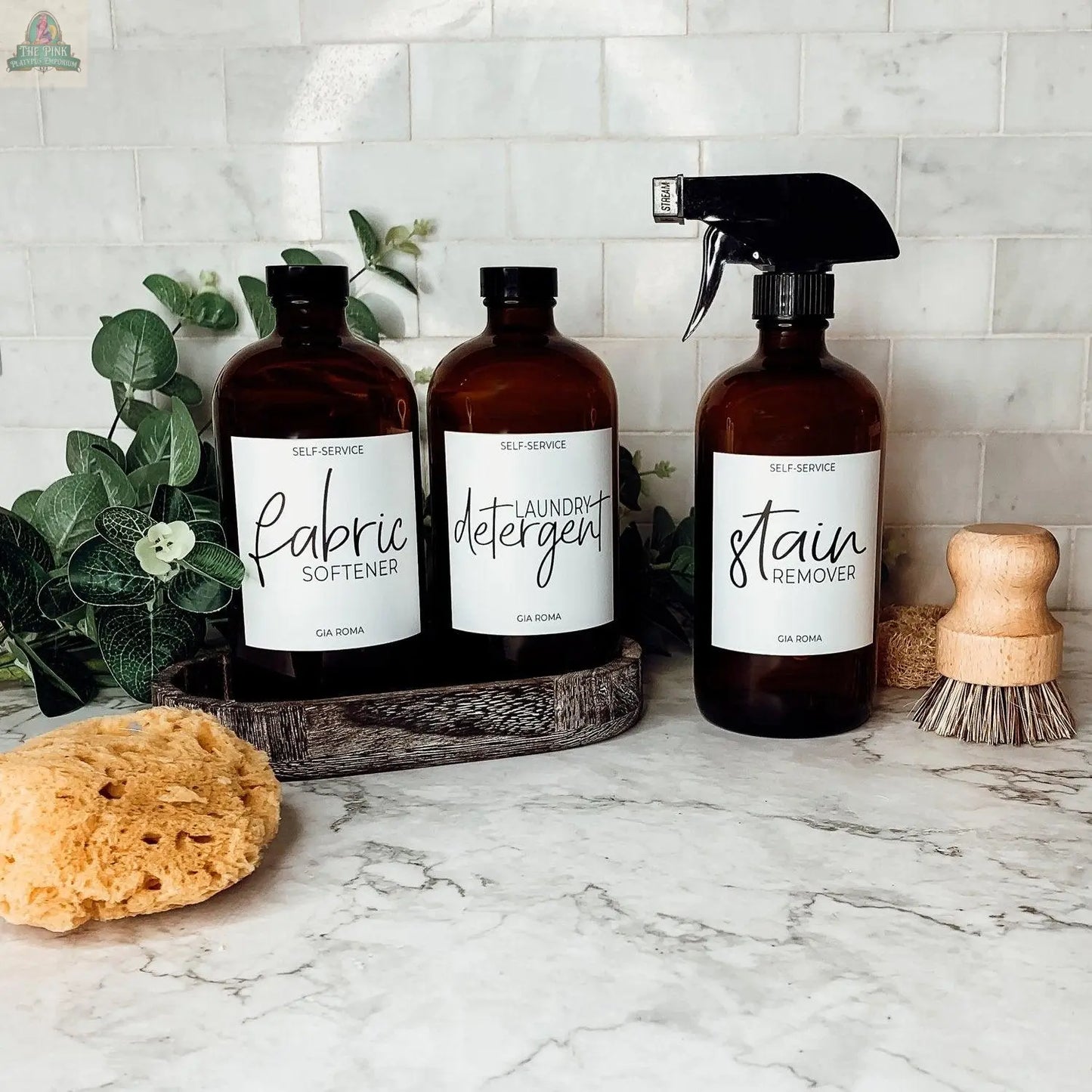Three 16oz Glass Amber | White Label bottles, labeled fabric softener, laundry detergent, and stain remover, sit on a countertop with a scrub brush, yellow sponge, greenery, and tile backsplash. Each bottle has a waterproof label.