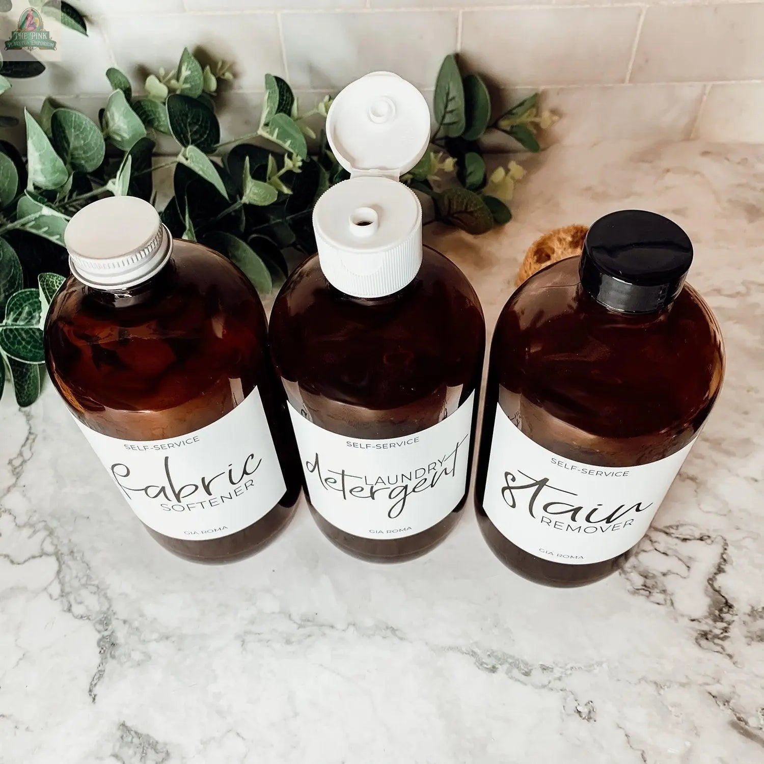 Three 16oz Glass Amber | White Label bottles—marked “Fabric Softener,” “Laundry Detergent,” and “Stain Remover”—are displayed on a marble countertop with waterproof labels, the detergent bottle open and greenery in the background.