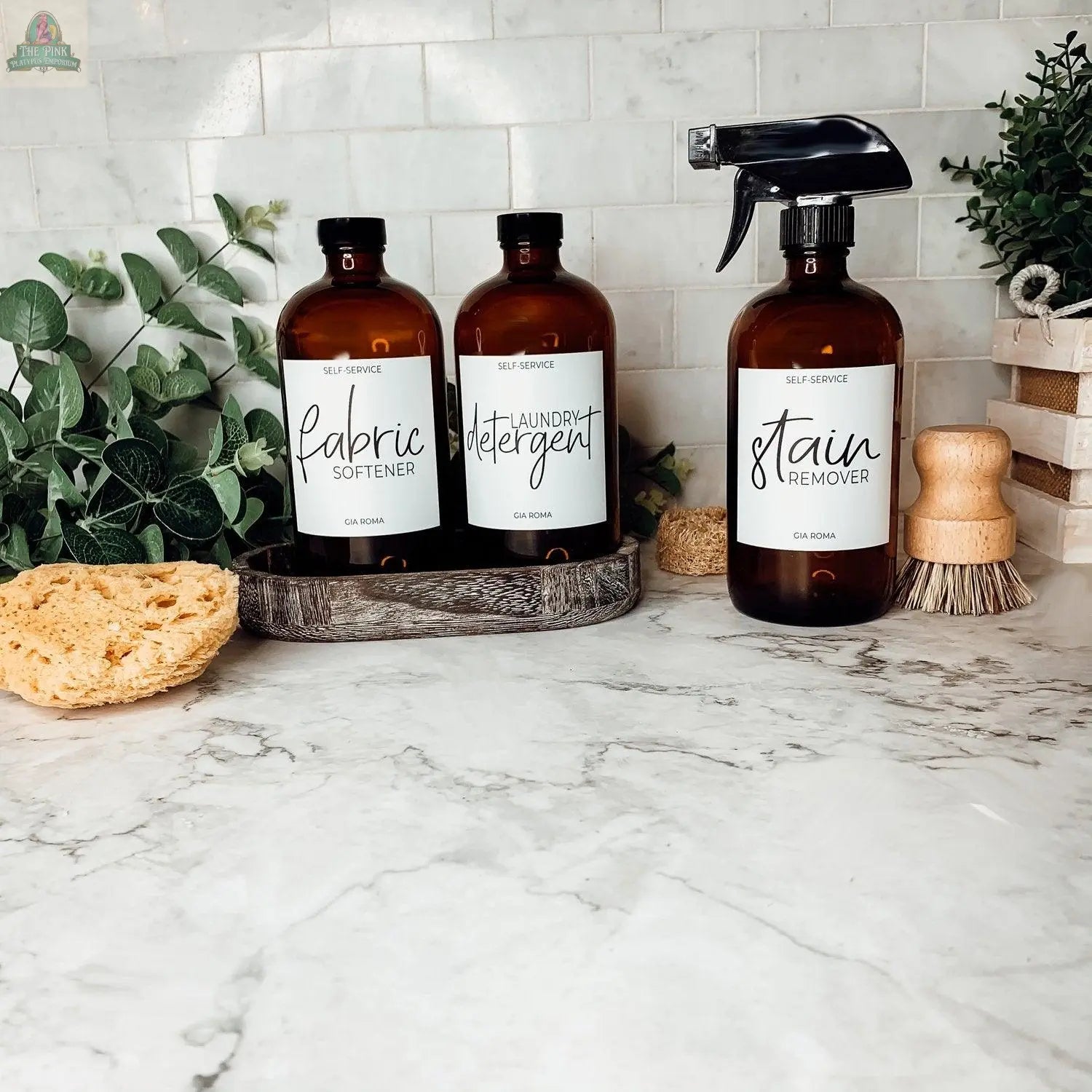 Three 16oz Glass Amber | White Label bottles sit on a marble countertop labeled fabric softener, laundry detergent, and stain remover, surrounded by brushes, a sponge, and green plants against a tiled backsplash.