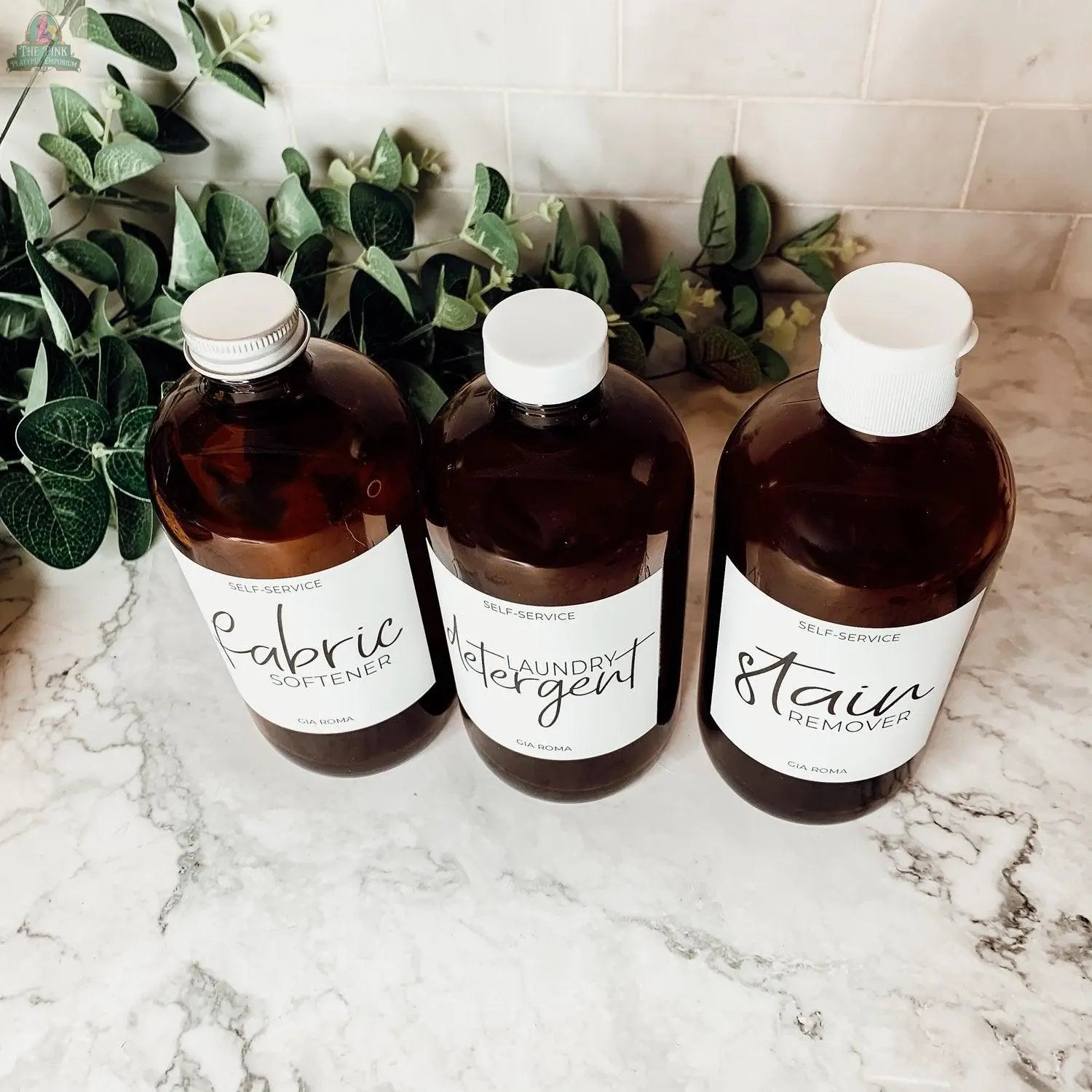 Three 16oz Glass Amber | White Label bottles labeled Fabric Softener, Laundry Detergent, and Stain Remover sit on a marble countertop with green leaves and white tile in the background.