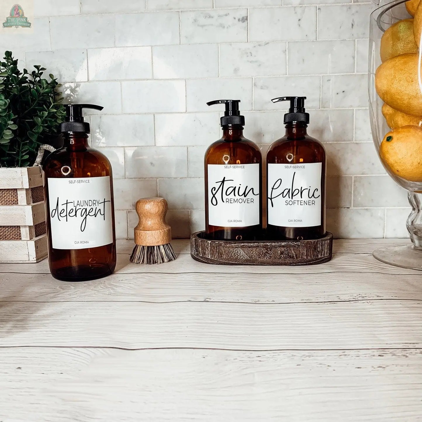 Three 16oz Glass Amber | White Label bottles—labeled “laundry detergent,” “stain remover,” and “fabric softener”—are displayed on a white wooden counter with a brush, potted plant, and a vase of lemons.