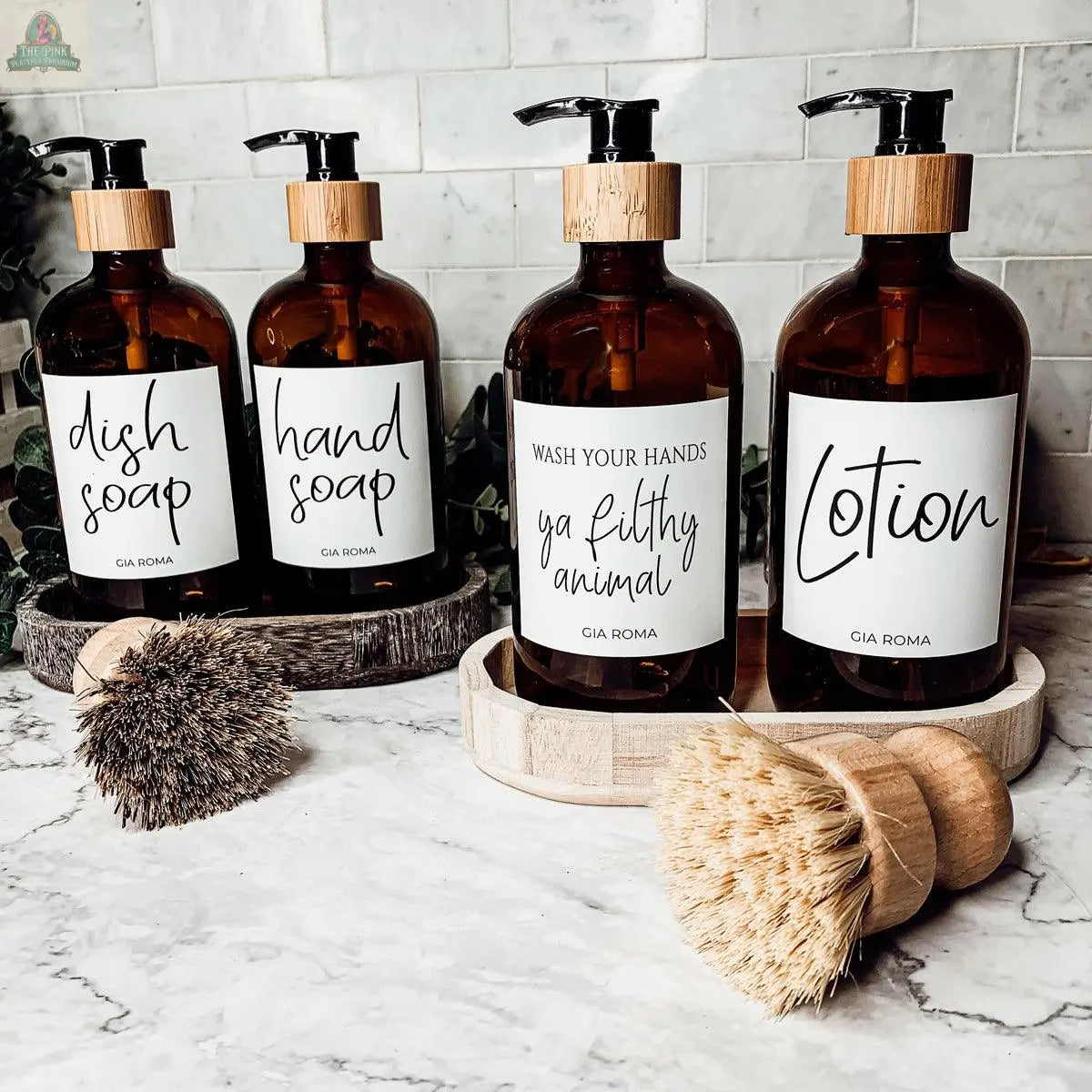 Four 16oz Glass Amber | White Label bottles—labeled dish soap, hand soap, “wash your hands ya filthy animal,” and lotion—serve as soap dispensers on a marble countertop, with two wooden scrub brushes and a white tile backsplash.