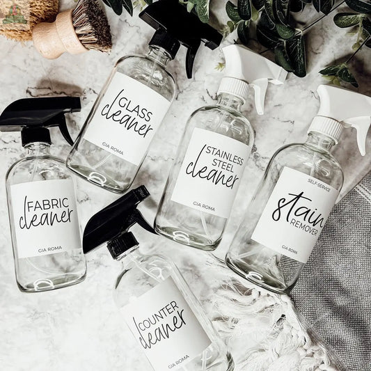 Five 16oz Glass Cleaning Bottles labeled fabric cleaner, glass cleaner, stainless steel cleaner, stain remover, and counter cleaner are displayed on marble with greenery, cleaning brushes, and glass dispensers beside them.