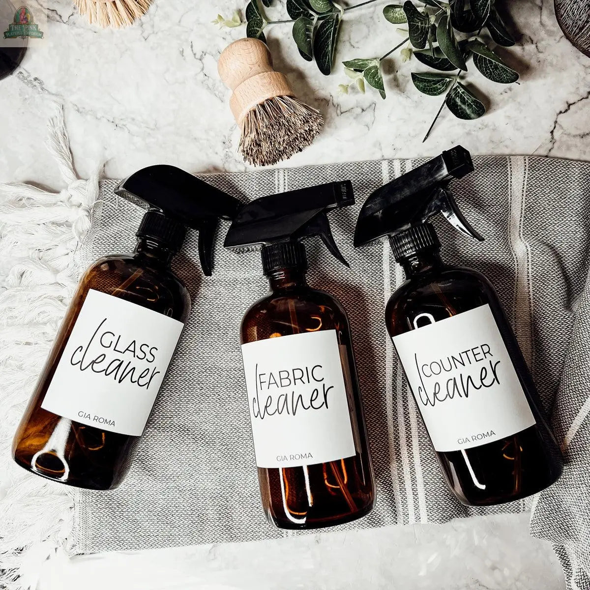 Three amber 16oz Glass Cleaning Bottles labeled Glass Cleaner, Fabric Cleaner, and Counter Cleaner rest on a striped cloth with a scrub brush and greenery—ideal refill bottles for your eco-friendly cleaning routine.