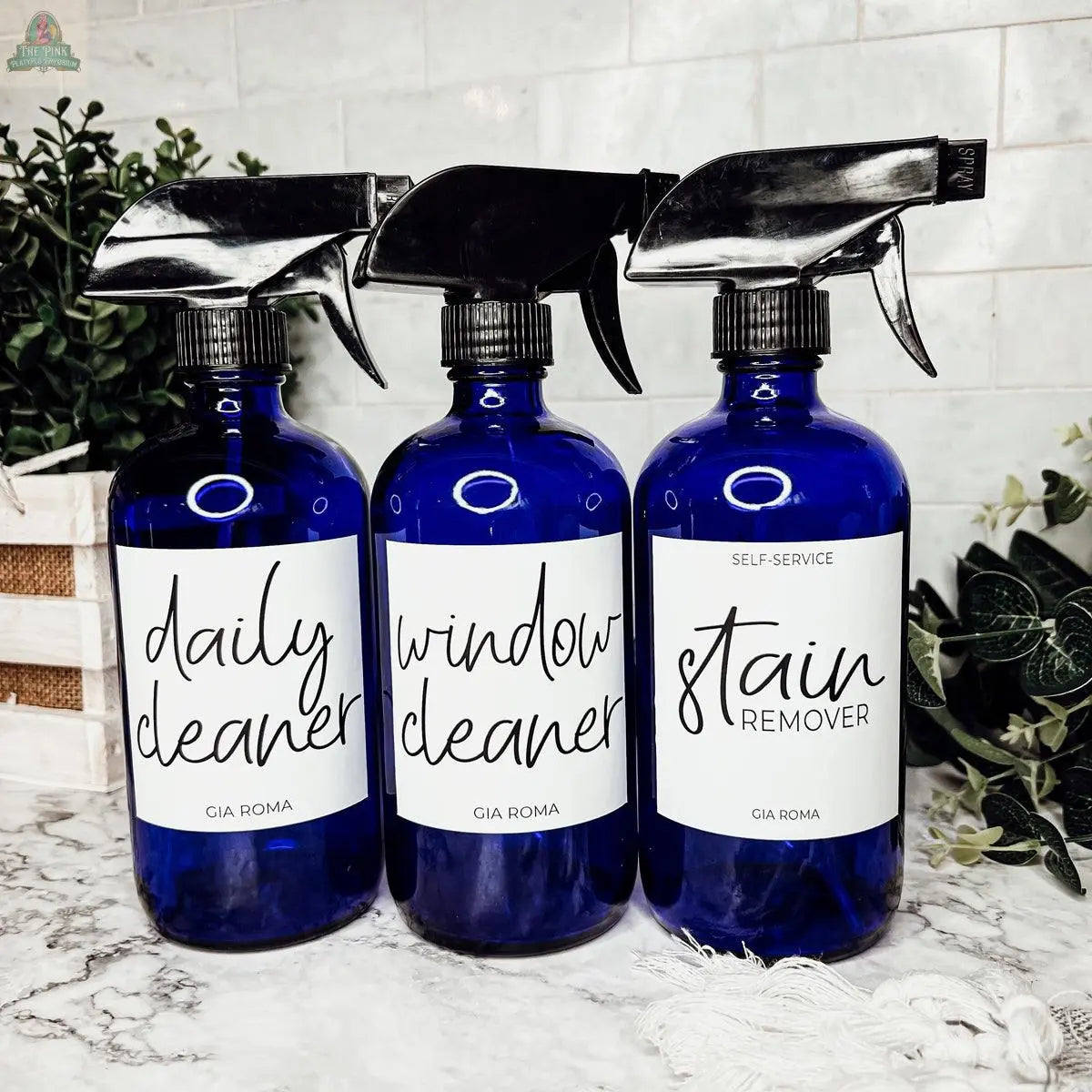 Three blue 16oz Glass Cleaning Bottles—labeled daily cleaner, window cleaner, and stain remover—sit on a marble countertop with green plants and white tile, making great refill bottles for any eco-friendly home.