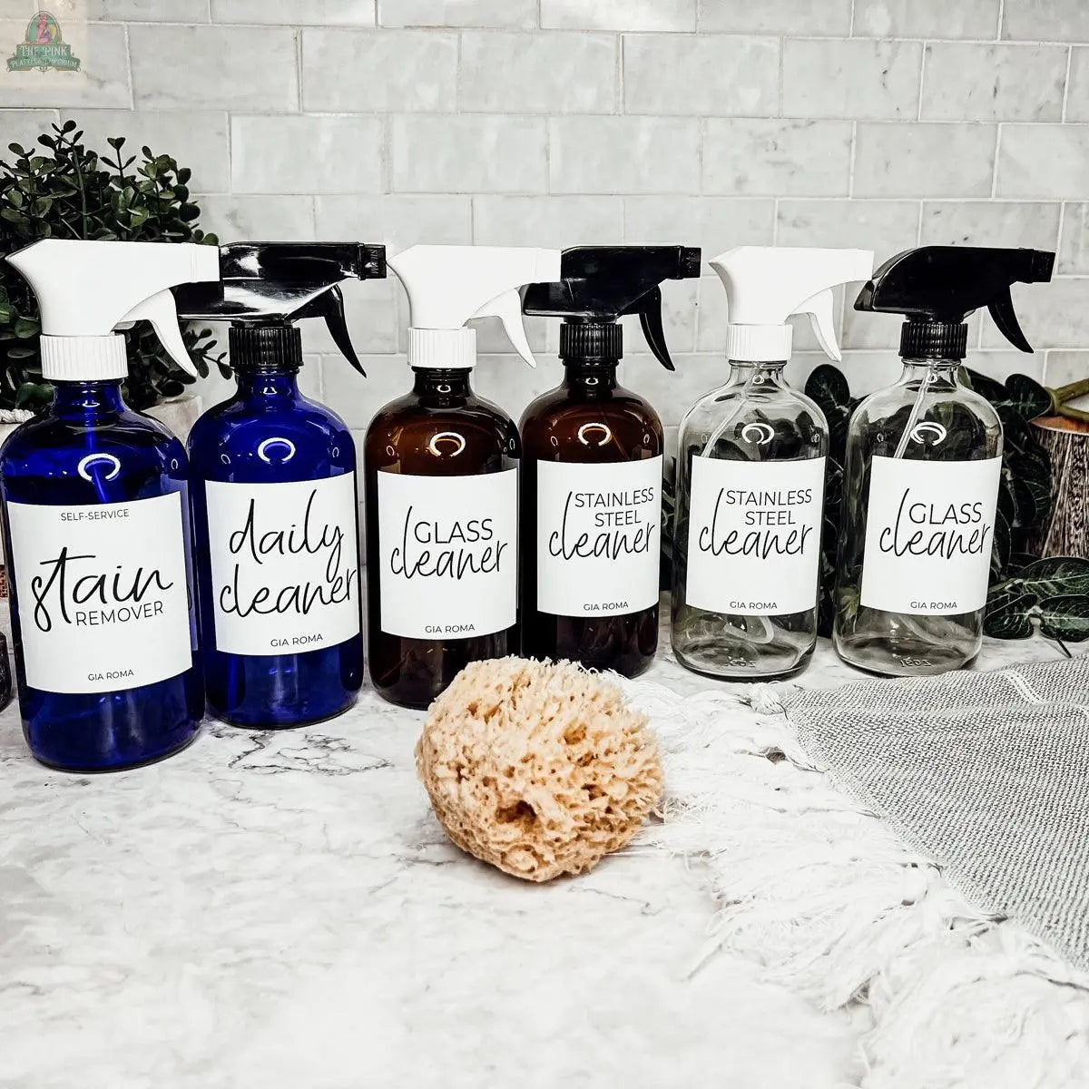 Six 16oz Glass Cleaning Bottles, each labeled for a different cleaner, are arranged on a marble countertop beside a natural sponge and greenery, with a white tiled backsplash in the background.