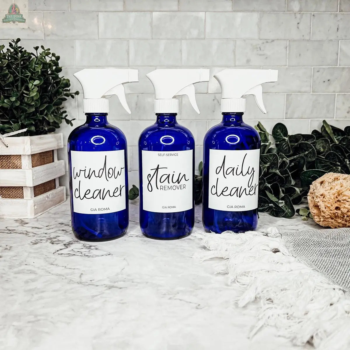 Three blue 16oz Glass Cleaning Bottles labeled window cleaner, stain remover, and daily cleaner sit on a marble countertop with greenery, a wooden crate, and a sponge—ideal refill bottles for your cleaning routine.