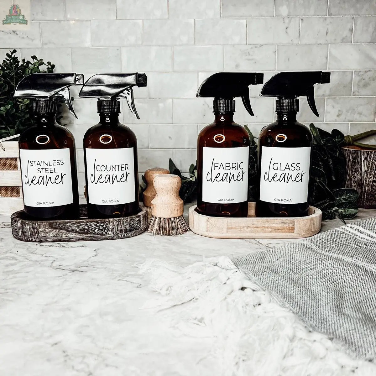 Four brown 16oz Glass Cleaning Bottles labeled Stainless Steel Cleaner, Counter Cleaner, Fabric Cleaner, and Glass Cleaner sit on a marble countertop with brushes and greenery, making stylish refill options for your home.
