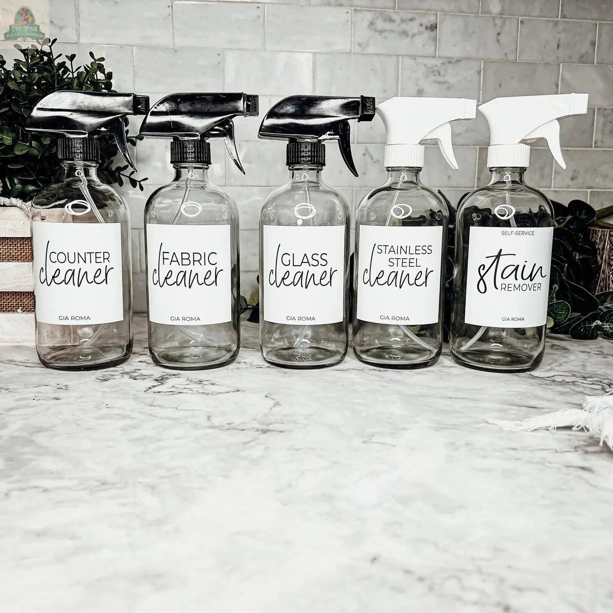 Five 16oz Glass Cleaning Bottles with black and white nozzles, each labeled for counter, fabric, glass, stainless steel, and stain remover, are displayed on a marble counter—ideal refill bottles for every cleaning need.