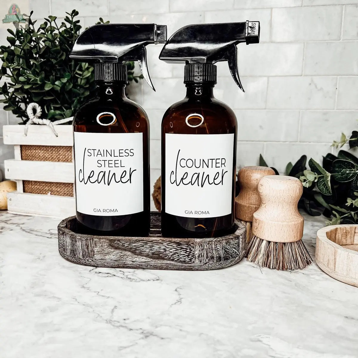 Two 16oz Glass Cleaning Bottles labeled Stainless Steel Cleaner and Counter Cleaner sit in a wooden tray on a marble countertop with cleaning brushes, greenery, and white baskets.