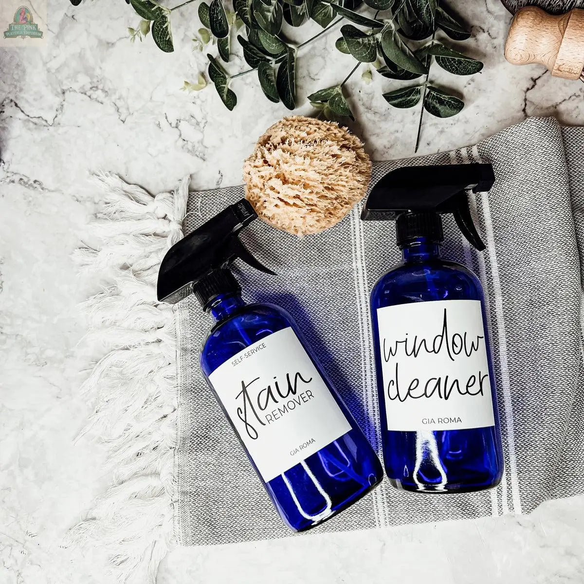Two blue 16oz Glass Cleaning Bottles labeled "Stain Remover" and "Window Cleaner" sit on a gray striped towel with a natural sponge and green leaves on marble. Ideal as refill bottles for your cleaning routine.