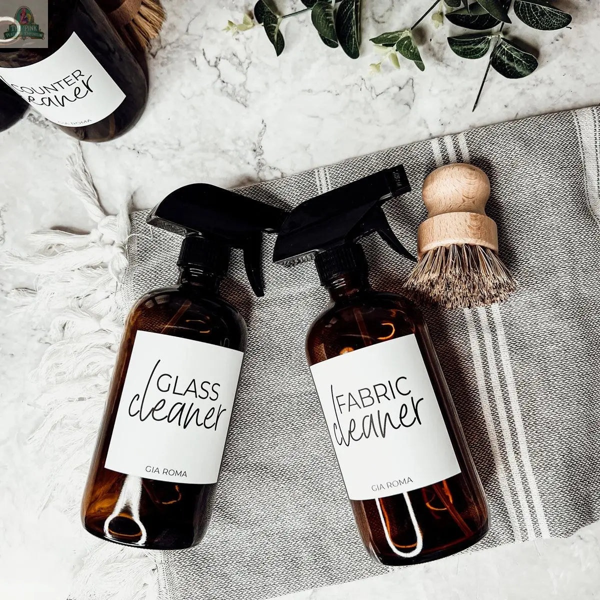 Two 16oz Glass Cleaning Bottles labeled Glass Cleaner and Fabric Cleaner rest on a gray striped cloth beside a wooden scrub brush, while a leafy plant and another glass dispenser appear in the background.