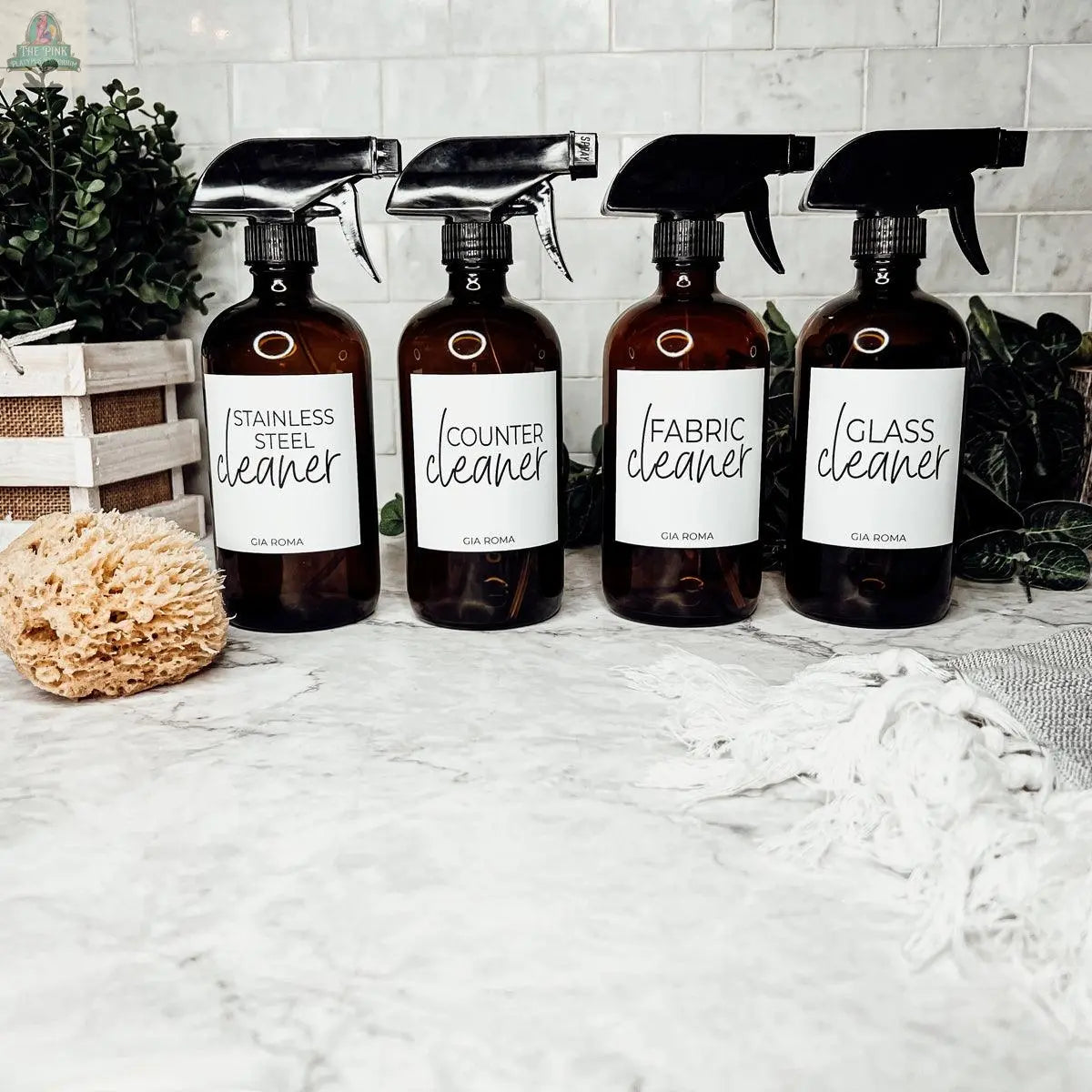 Four brown 16oz Glass Cleaning Bottles labeled stainless steel, counter, fabric, and glass cleaner sit on a marble countertop with a sponge, plants, and white towel—perfect refill bottles to elevate your cleaning routine.