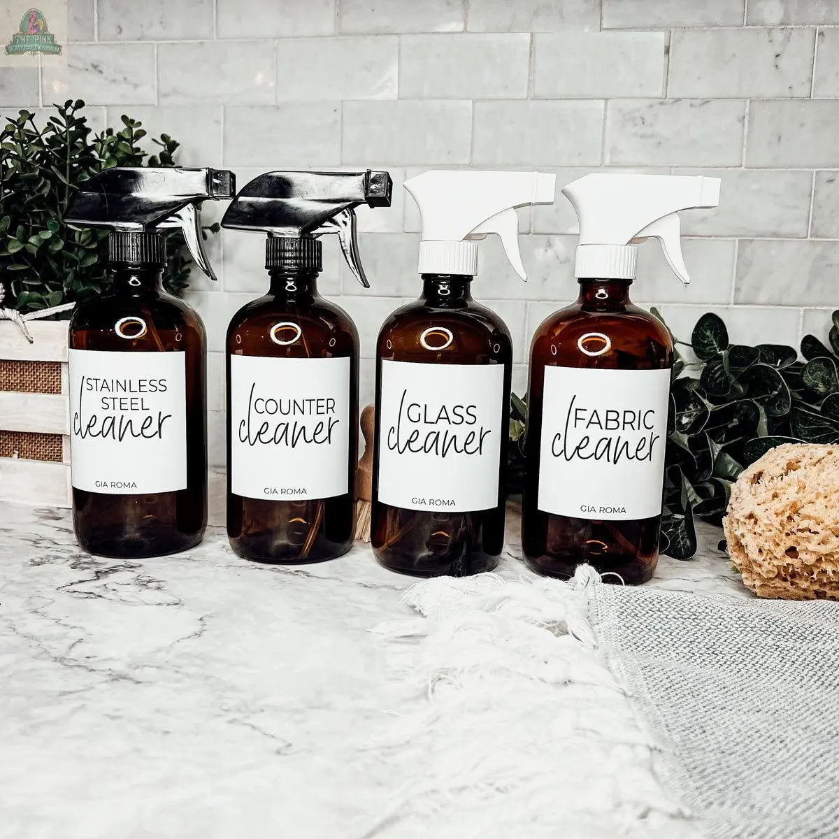 Four 16oz Glass Cleaning Bottles labeled Stainless Steel Cleaner, Counter Cleaner, Glass Cleaner, and Fabric Cleaner are arranged on a marble counter with plants and cloths, making them perfect refill bottles for any home.