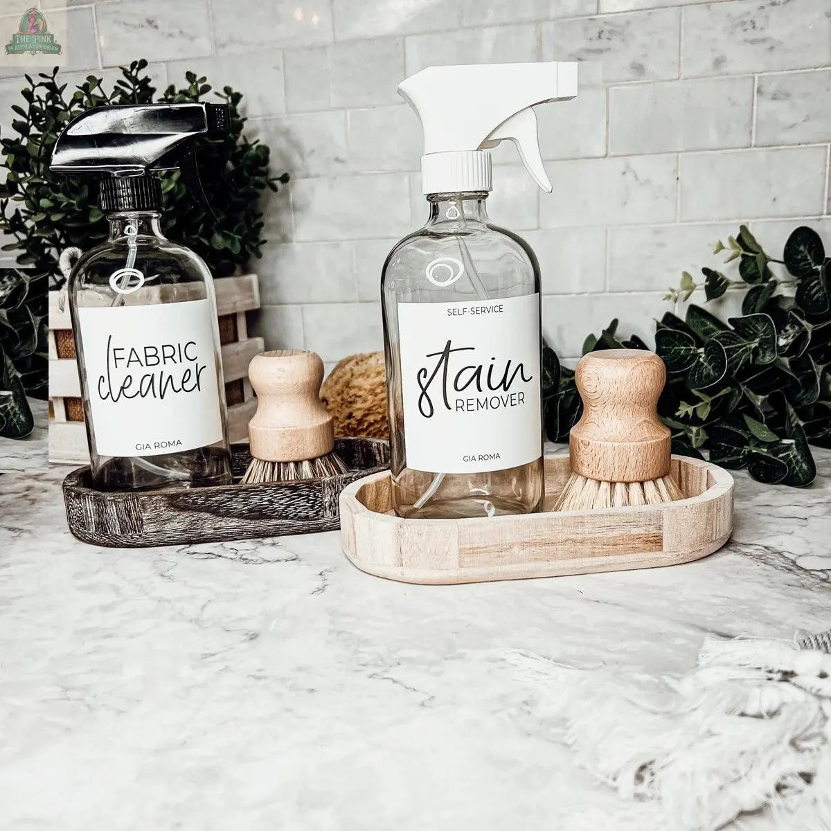 Two 16oz Glass Cleaning Bottles labeled Fabric Cleaner and Stain Remover rest in wooden trays on a marble countertop, surrounded by scrubbing brushes and green plants, adding a stylish touch to your cleaning routine.